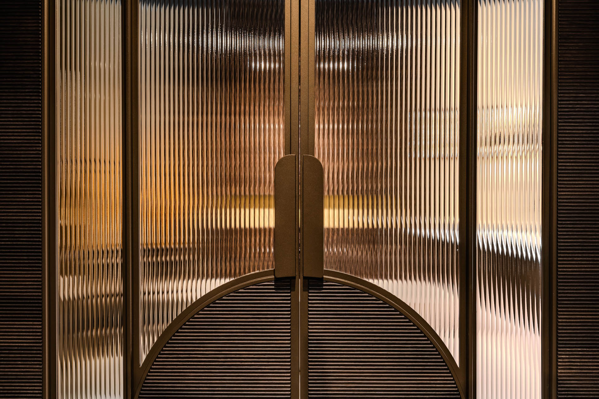 Close-up van bronskleurige stalen deur met ribbelglas, ronde panelen en bronzen handgrepen.