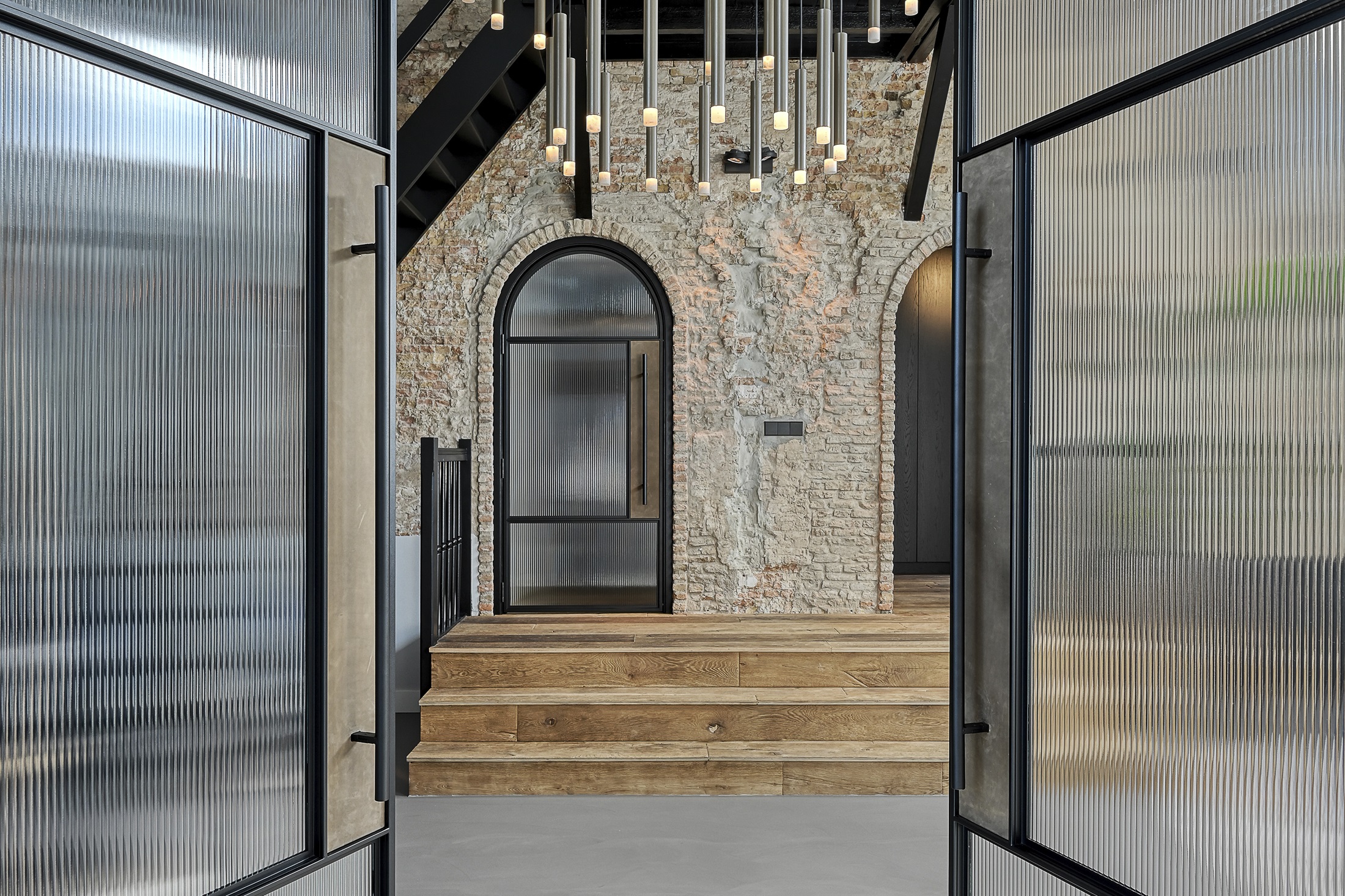 Zwarte stalen deuren met geribbeld glas en een boogvorm, gecombineerd met een rustieke bakstenen muur en moderne verlichting.