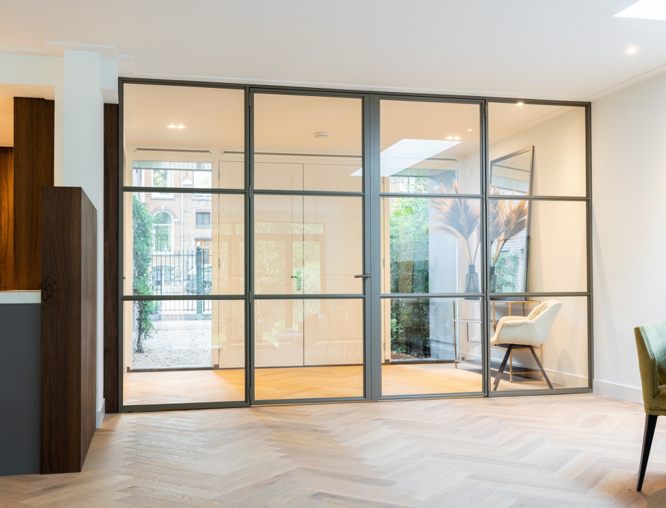 Elegante Anodic Bronze stalen roomdivider met glas, die een lichte ruimte met visgraatvloer en tuinzicht scheidt.