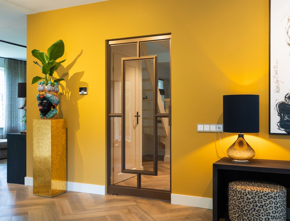 Bronzen stalen taatsdeur met glas in een modern interieur met gele accentmuur en visgraatvloer.