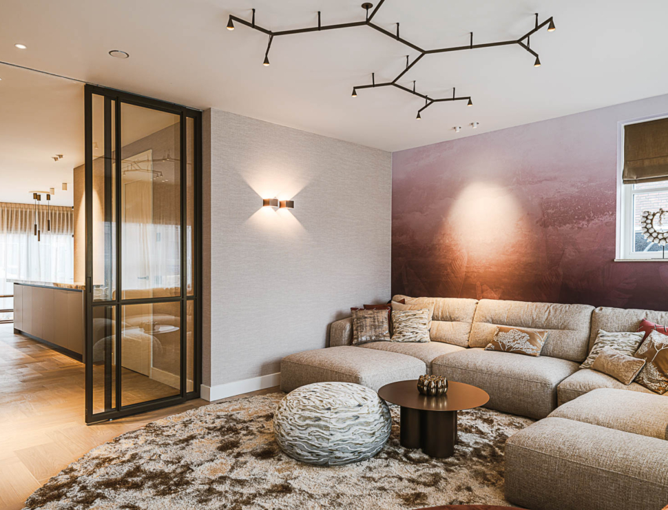 Gezellige woonkamer met beige hoekbank, glazen stalen deur en warme verlichting op een textuurrijke muur.