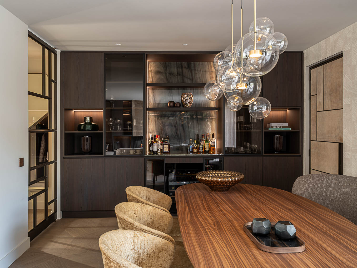 Luxe eetkamer met maatwerk barmeubel in donker hout, glazen stalen deur en opvallende designlamp met glazen bollen.