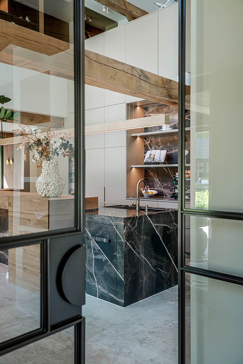 Doorkijk naar luxe keuken met marmeren werkblad en stalen taatsdeur met glas.
