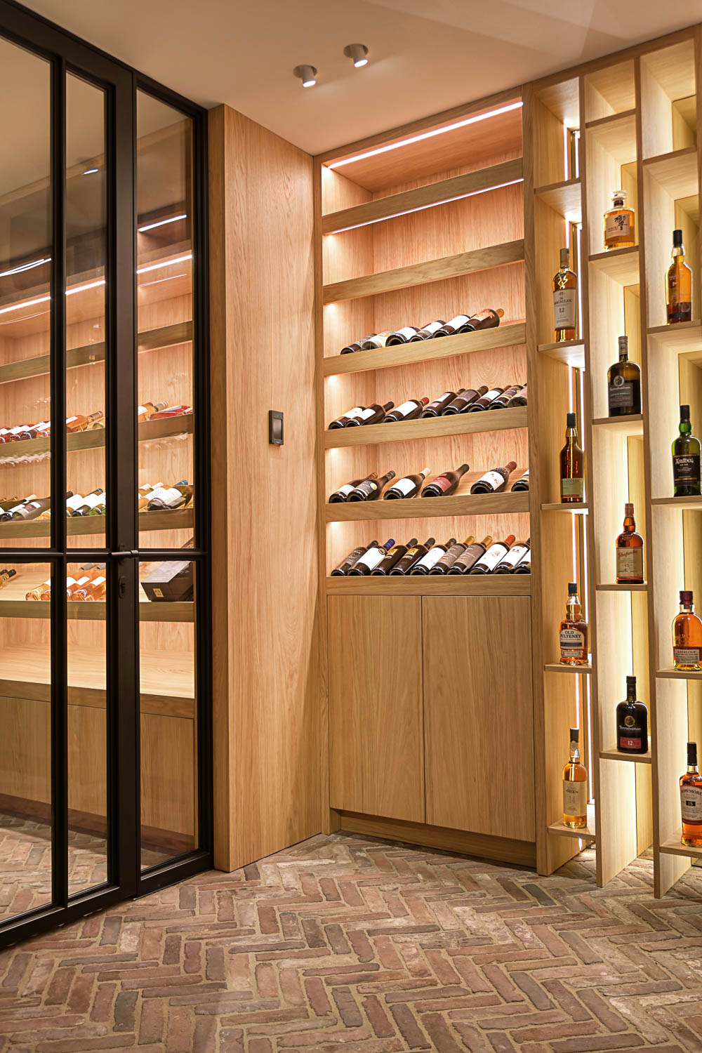 Zwarte stalen wijnkastdeur met glas in luxe wijnkelder met eikenhouten schappen en sfeerverlichting.