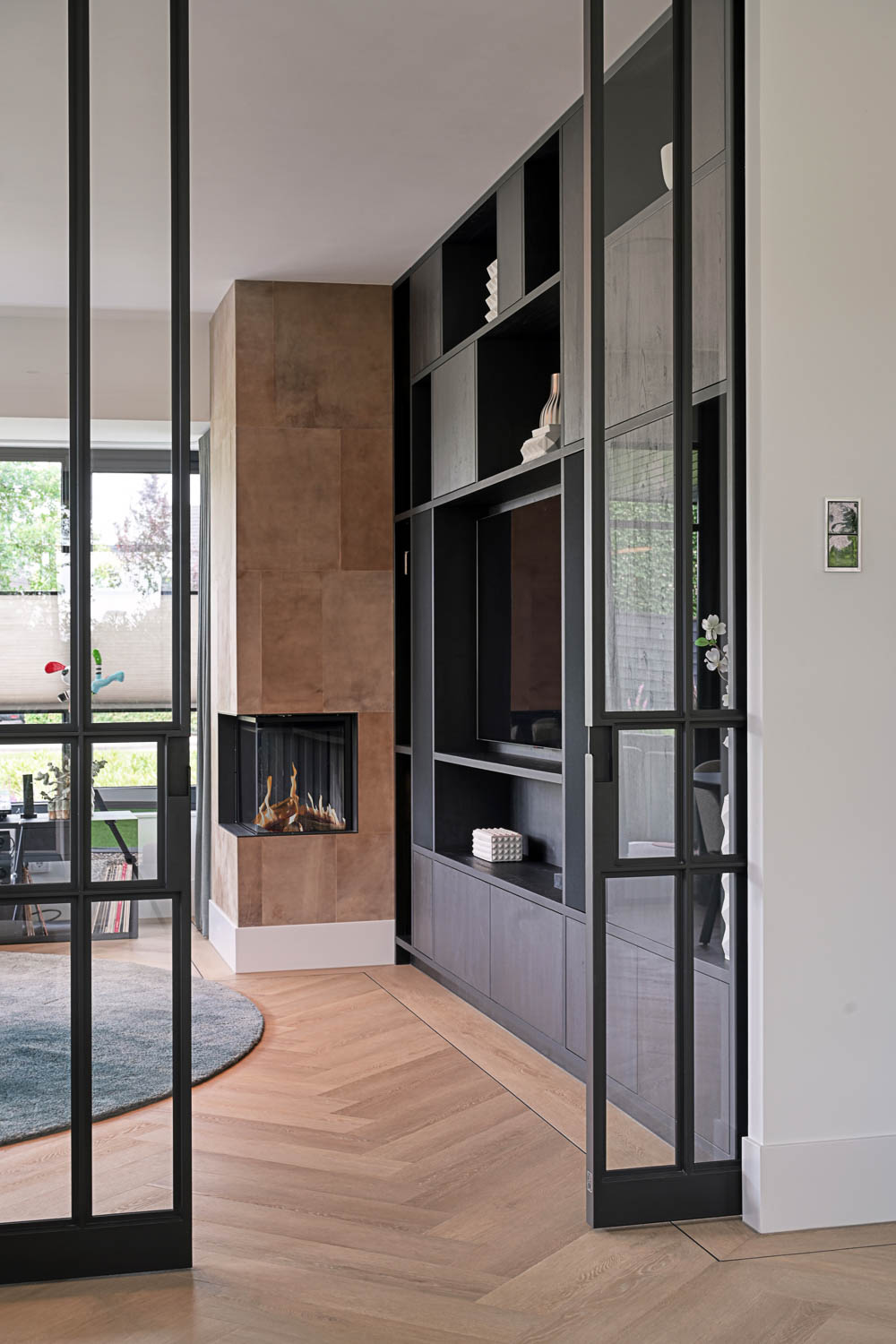 Zwarte maatwerk wandkast met open haard en stalen glasdeuren in stijlvol interieur met visgraatvloer.