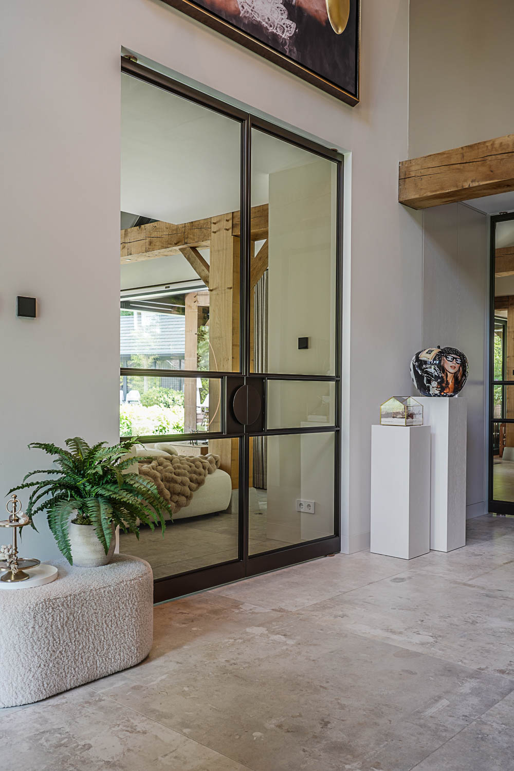 Zwarte stalen taatsdeur met glas in stijlvolle hal met kunstobject en beige natuurstenen vloer.