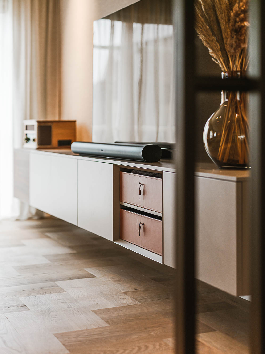 Minimalistisch zwevend tv-meubel met leren lades, een soundbar, stalen deur en decoratieve accessoires.